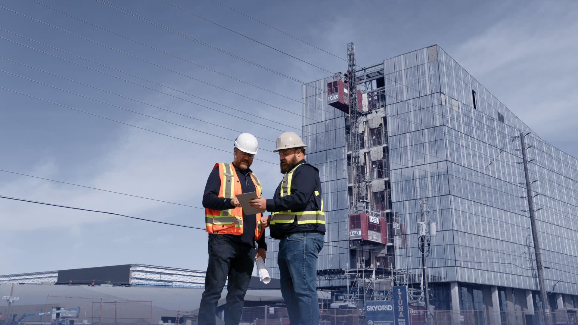 Two men in hard hats stand in front of a tall building using a tablet