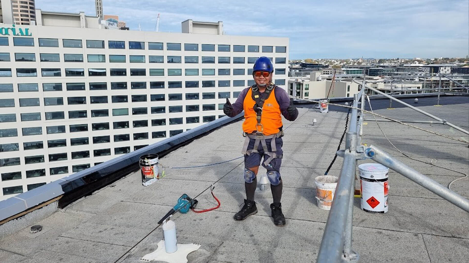 Construction worker smiling and giving thumbs up to the camera while standing on a roof top with safety gear on