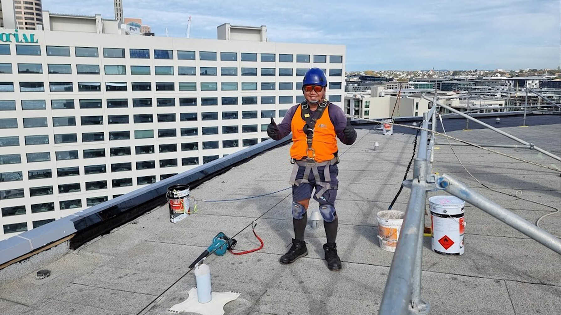 Construction worker smiling and giving thumbs up to the camera while standing on a roof top with safety gear on