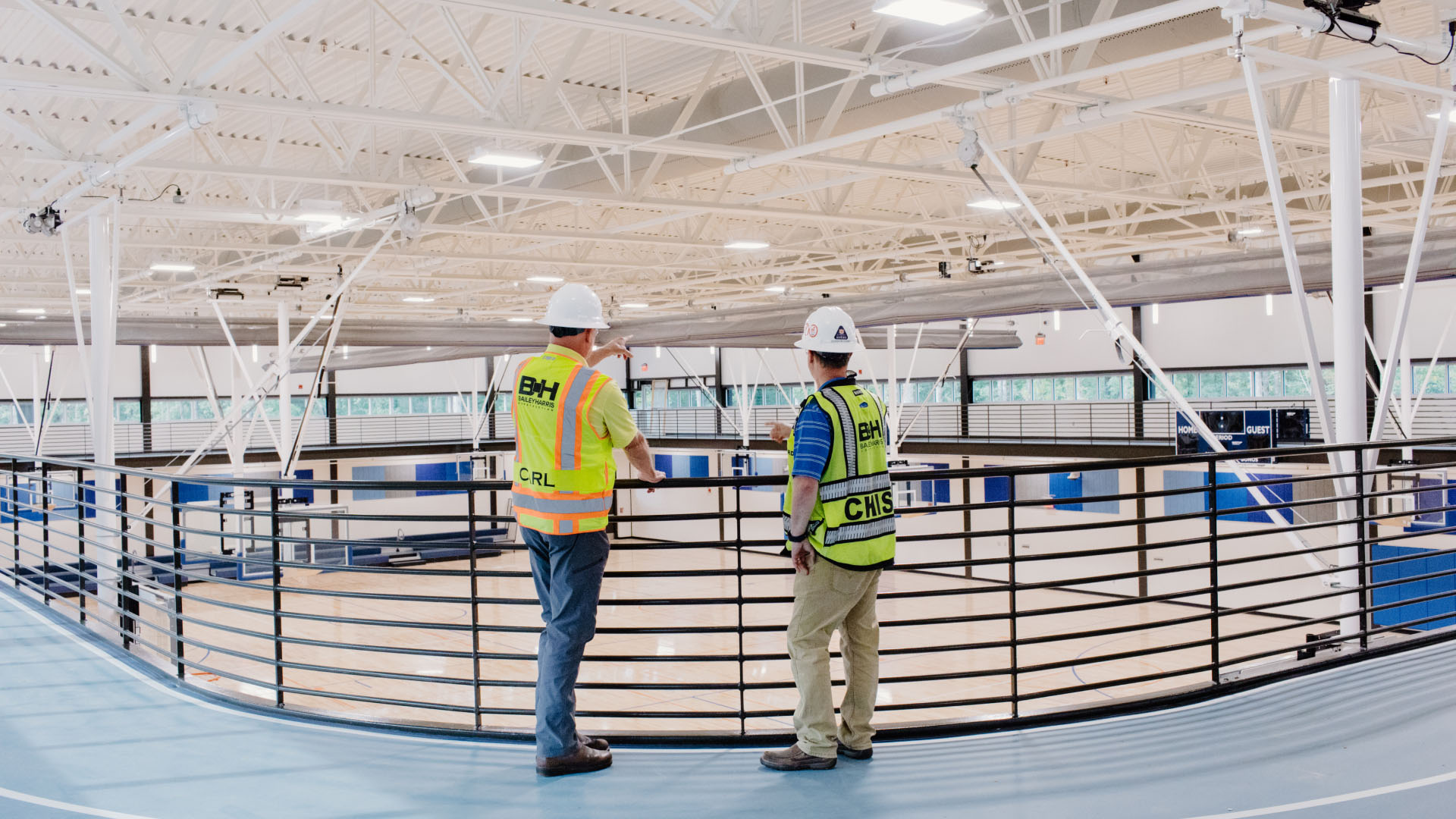 Two men in safety vests observe a basketball court from a balcony