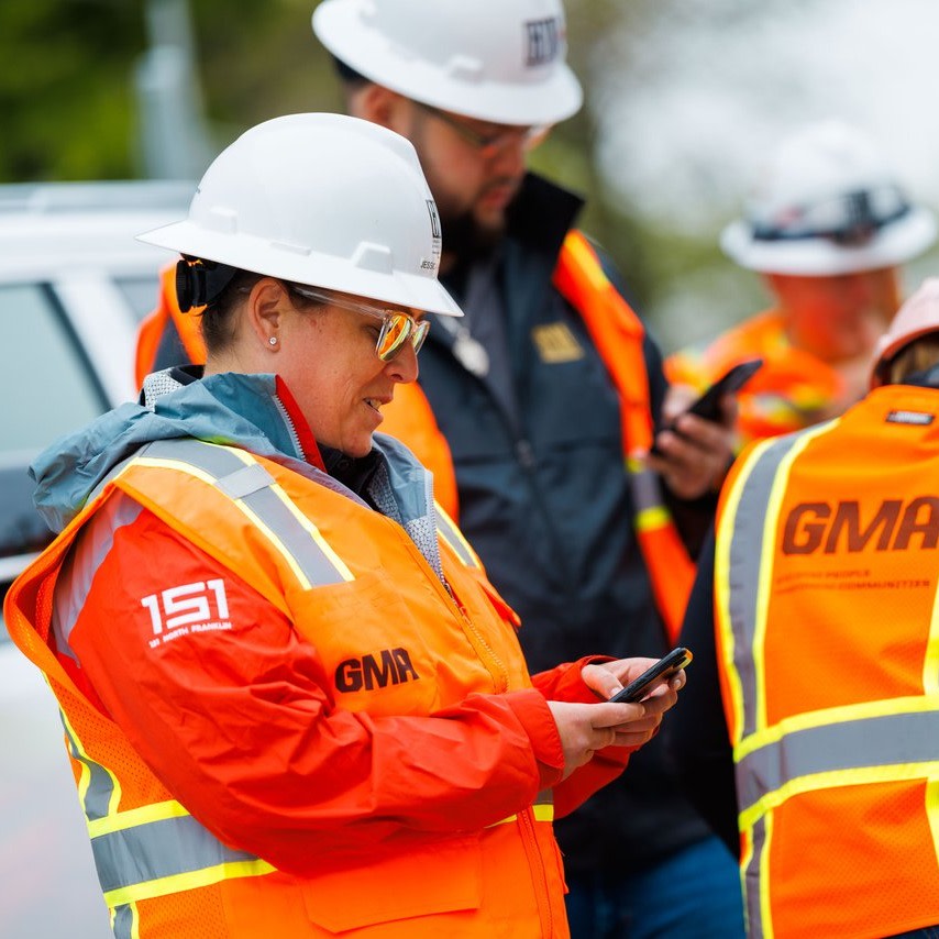 Construction workers in orange safety vests and white helmets using smartphones outdoors.