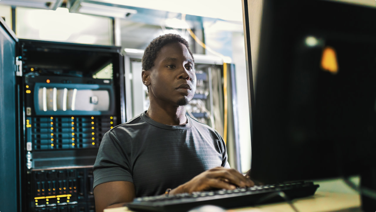Man working on a computer in a server room with network equipment behind him.