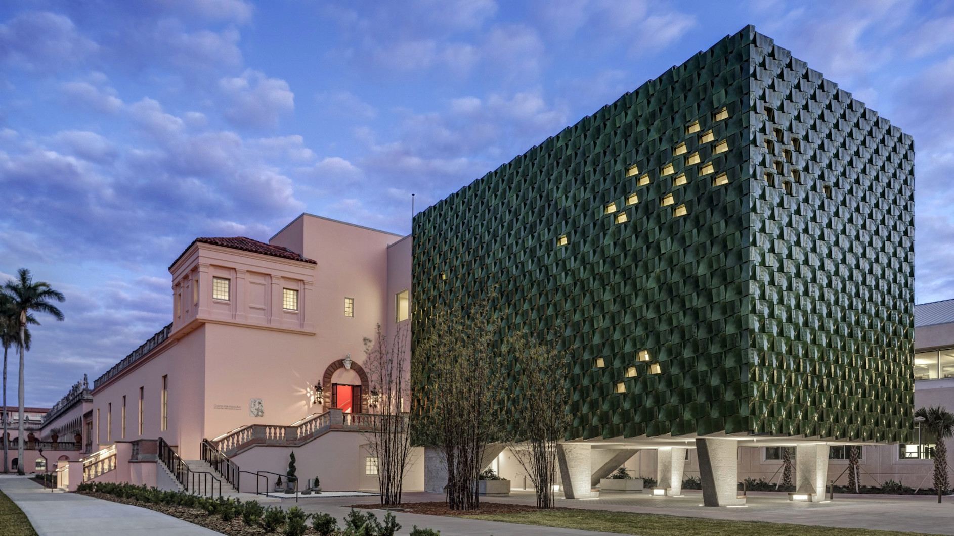 Modern green-tiled building addition next to historic pink Mediterranean-style structure under twilight sky with illuminated windows