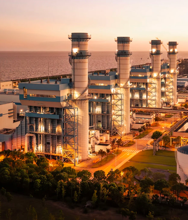Illuminated power plant with four tall smokestacks at sunset by the ocean.