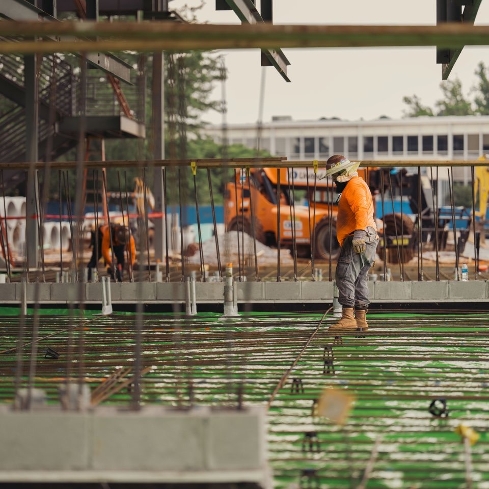 Construction workers in orange gear standing on a rebar grid at a site.