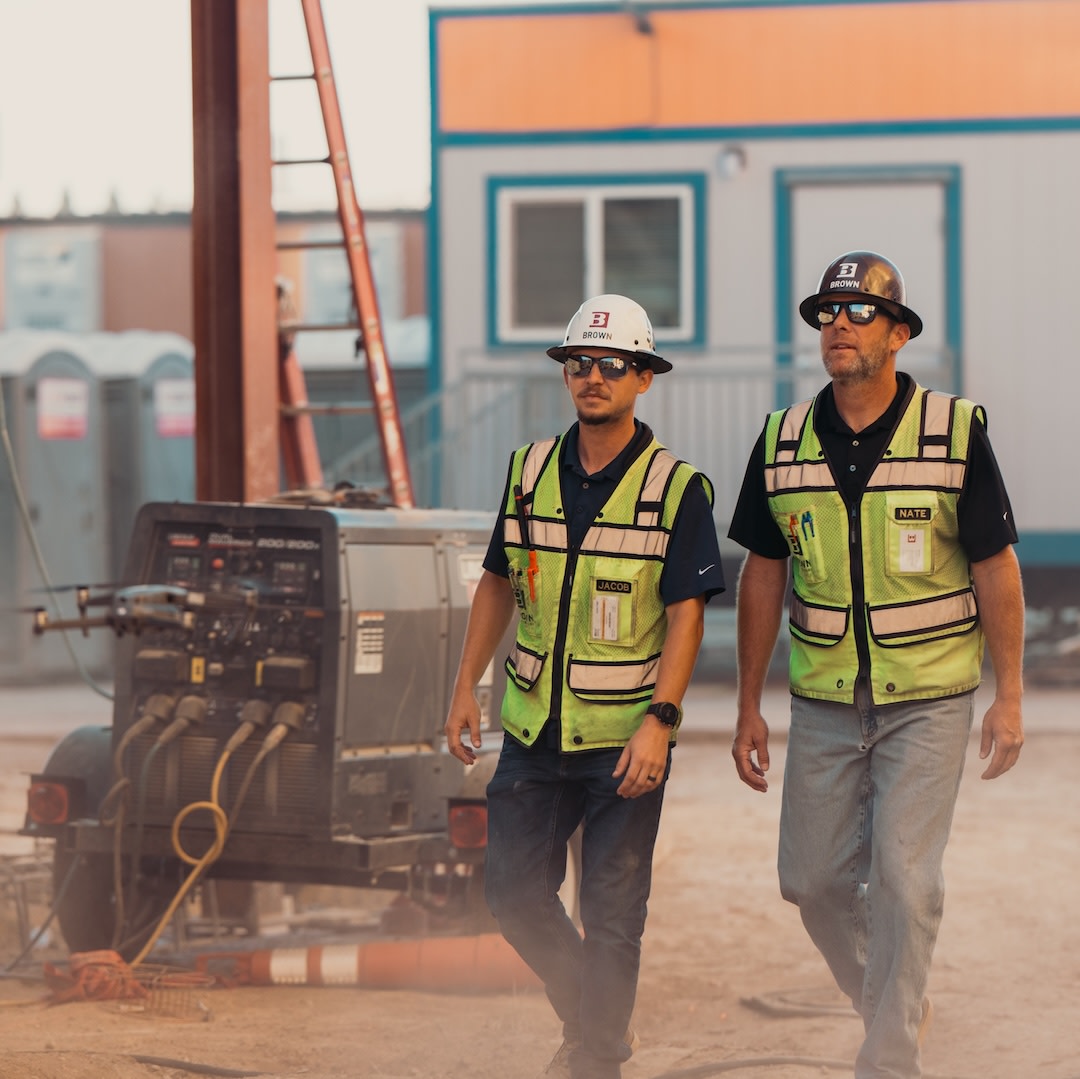 Two construction workers in safety vests and hard hats walking on a job site with equipment and buildings.