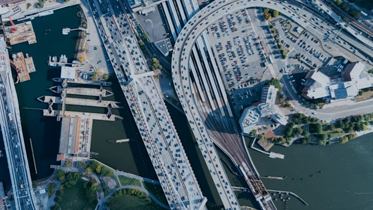 Aerial view of urban harbor with ships, parking lots, highways, and waterfront infrastructure.