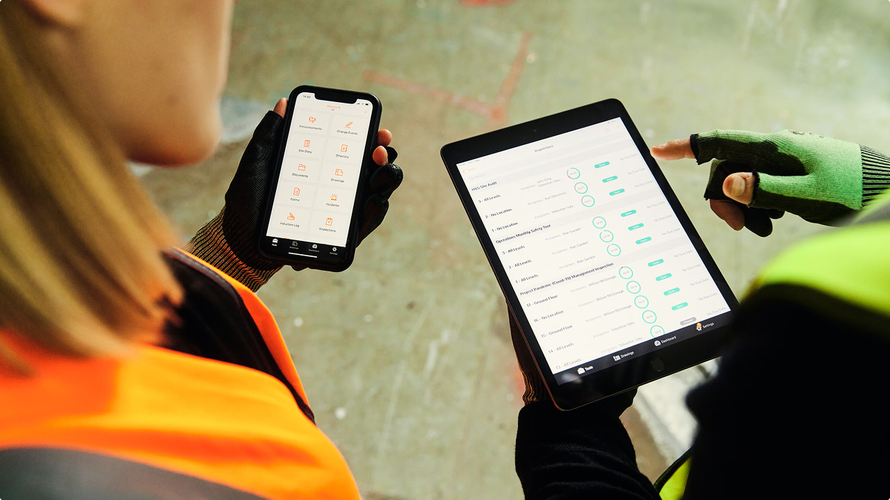 Two construction workers in safety vests using a smartphone and tablet.