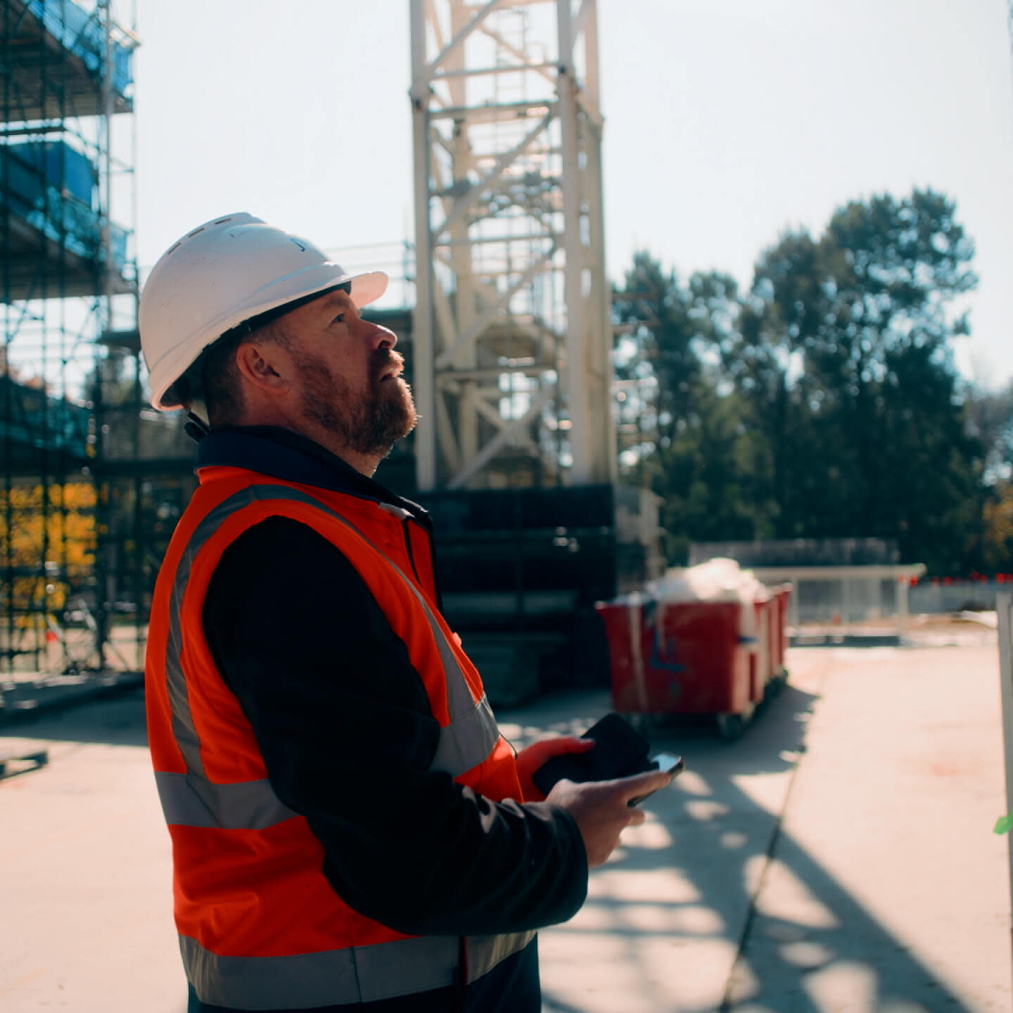 a contractor in a hard hat and vest holding a tablet while looking up at the construction site
