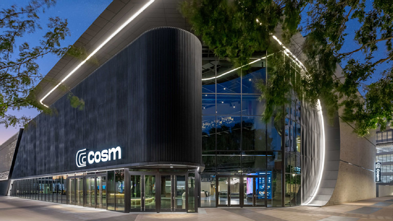 Modern building with curved glass facade and illuminated "cosm" sign at dusk
