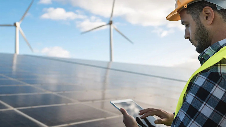 construction worker inspecting solar panels with an ipad with wind turbine in background