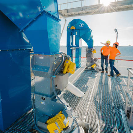 Two workers in orange shirts beside a machine, having a conversation and overseeing its operation