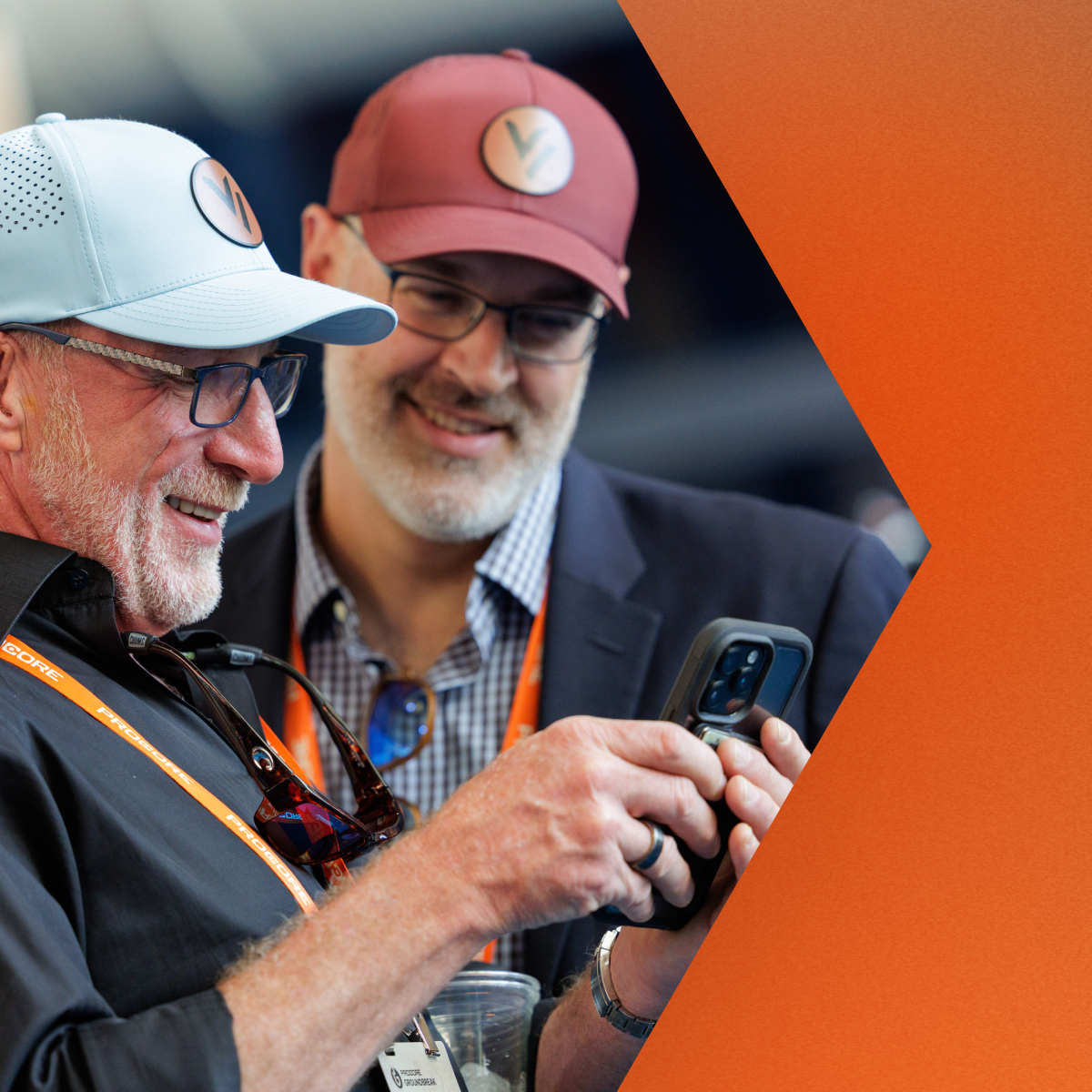 Two men wearing caps and glasses looking at a smartphone together outdoors.