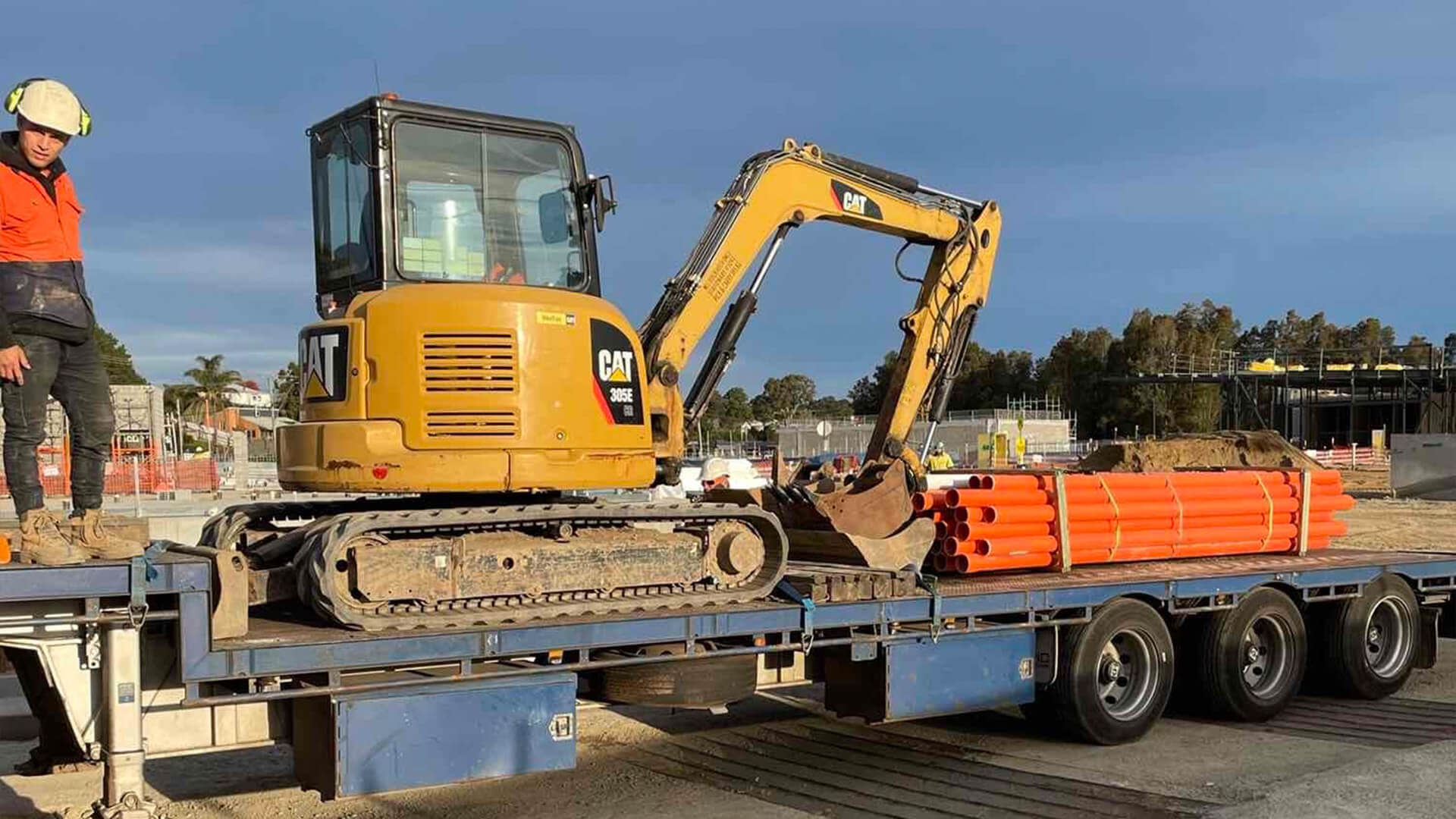 Subcontractor on top of a moving platform transporting a small excavator and pipes