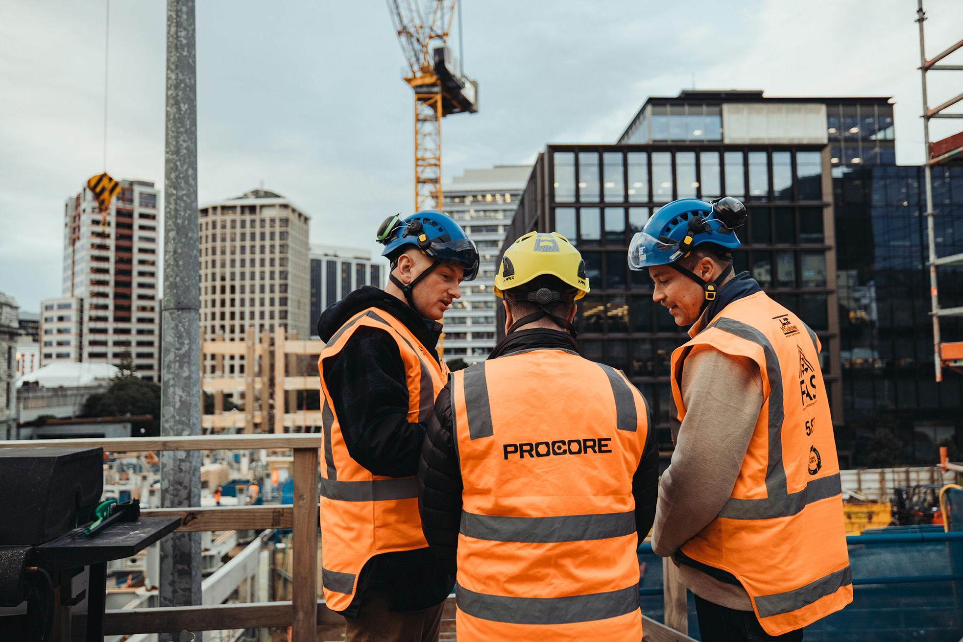 Three construction workers in orange vests and helmets discussing at a site.