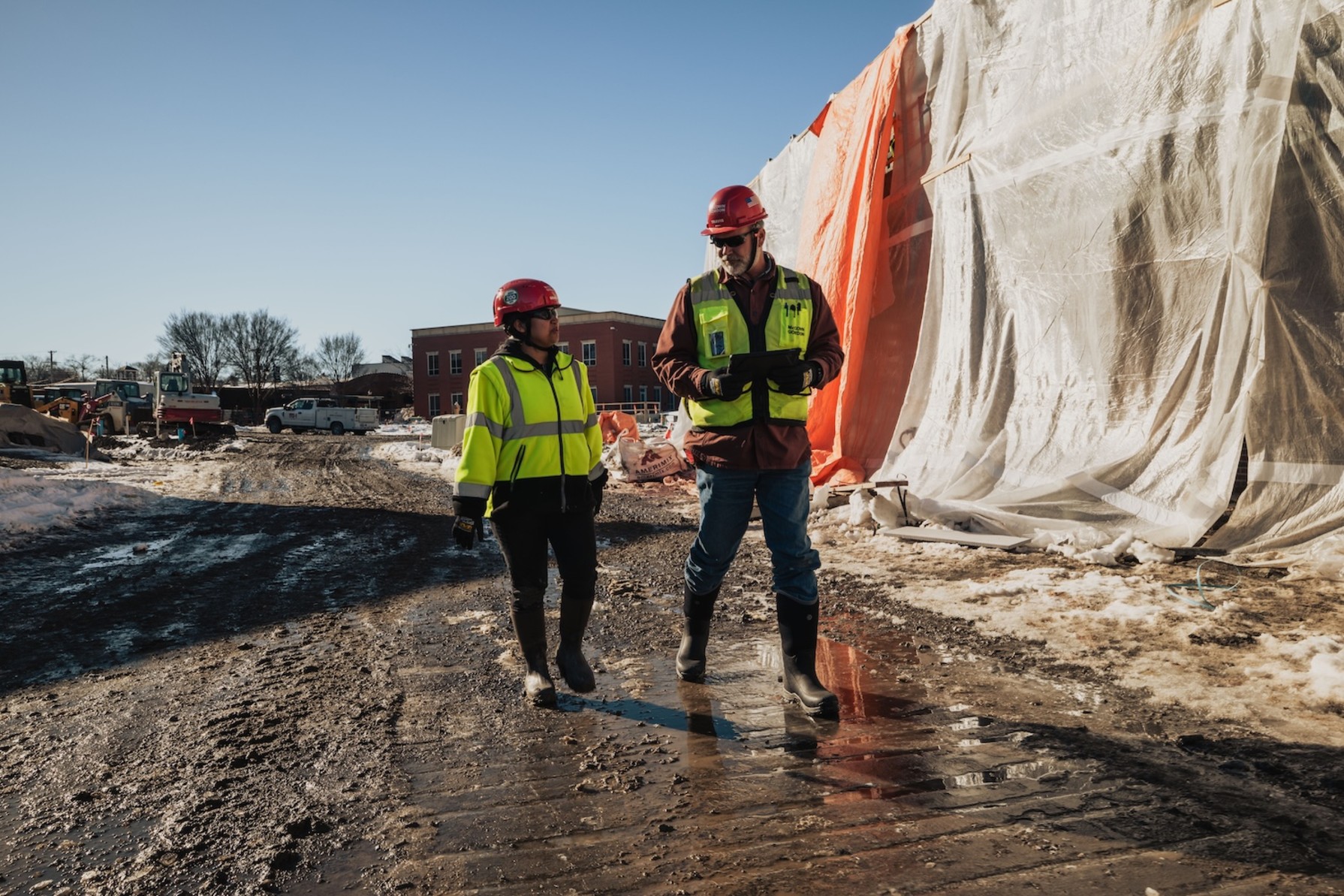 Two construction workers in safety gear walking on muddy ground near large white and orange tarps at a job site.