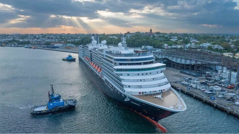 a large cruise ship in australia on cloudy day pulls out of port, there is a tug boat on one side and low density development on land.