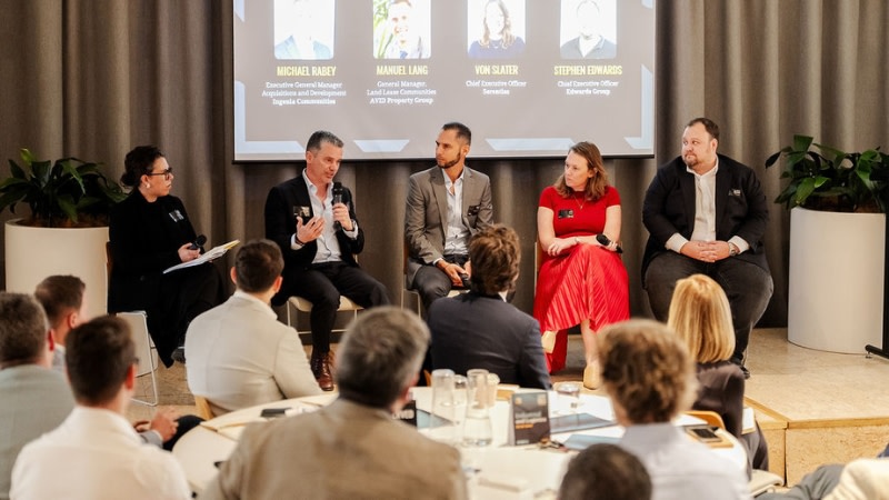five people on a panel talking on microphones in front of an audience in a modern conference room