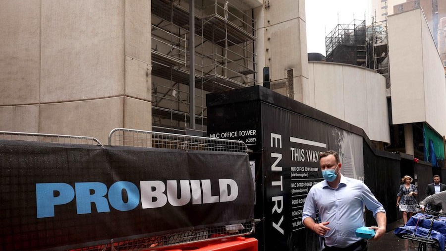 Man in mask walks past Probuild sign in city street.