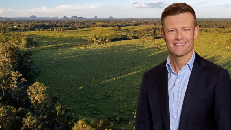 a man in a business suit smiles and is superimposed over a greenfield site in Rocksberg queensland with the glasshouse mountains behind them.