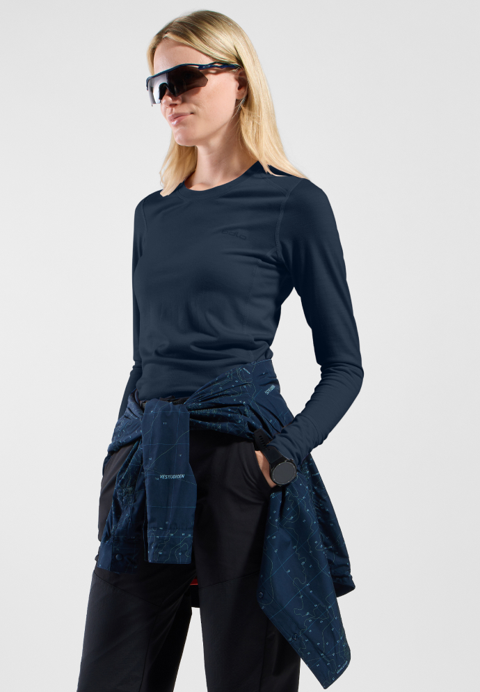 Blonde vrouw in een marineblauwe top met lange mouwen, een zwarte broek en een zonnebril, met een jasje met print om haar middel geknoopt.
