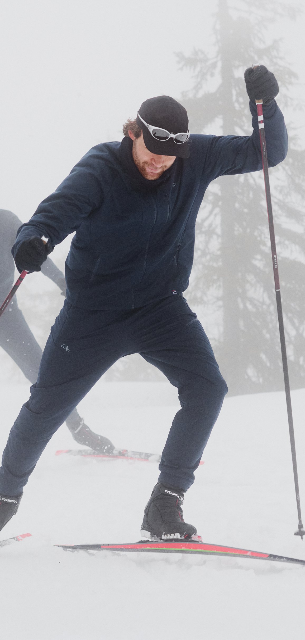 Zwei Langläufer gleiten durch den dichten Nebel eines verschneiten Waldes, gekleidet in dunkle Langlauf Sportbekleidung und Sonnenbrillen.