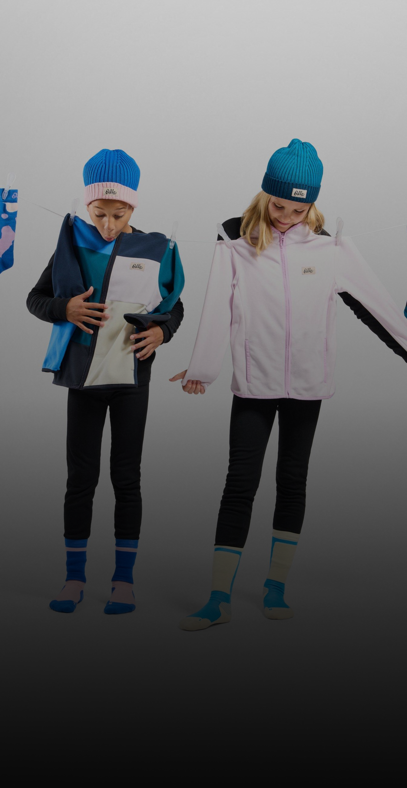 Twee kinderen met blauwe mutsen staan achter een waslijn met opgehangen sokken, met een blauwe jas en een roze jas in hun handen.