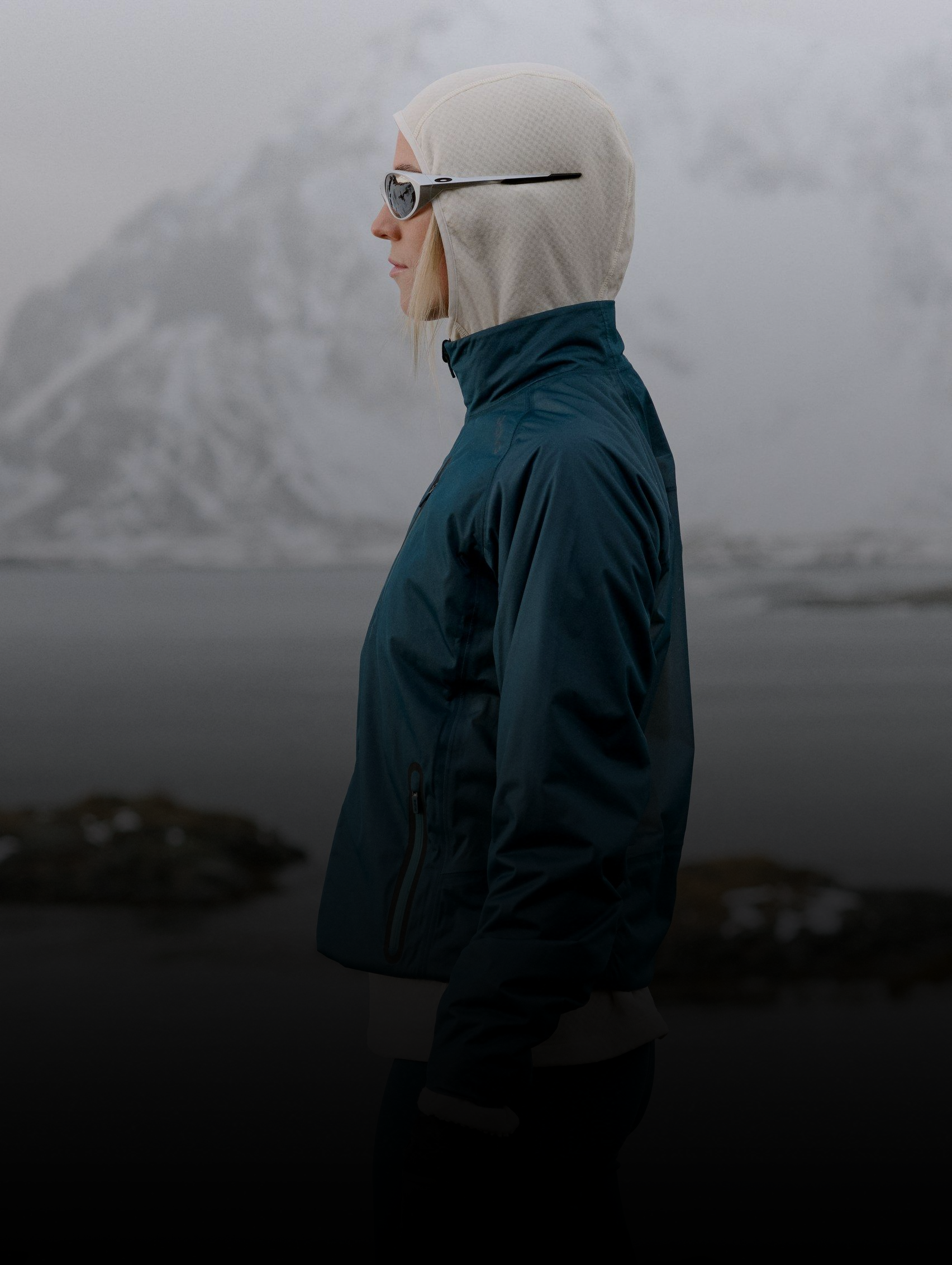 Frau in einer blaugrünen Dual Dry wasserdichten Jacke, schwarzer Weste und weißem Sturmhoodadjustiert ihre Sonnenbrille mit behandschuhten Händen in einer verschneiten Landschaft.