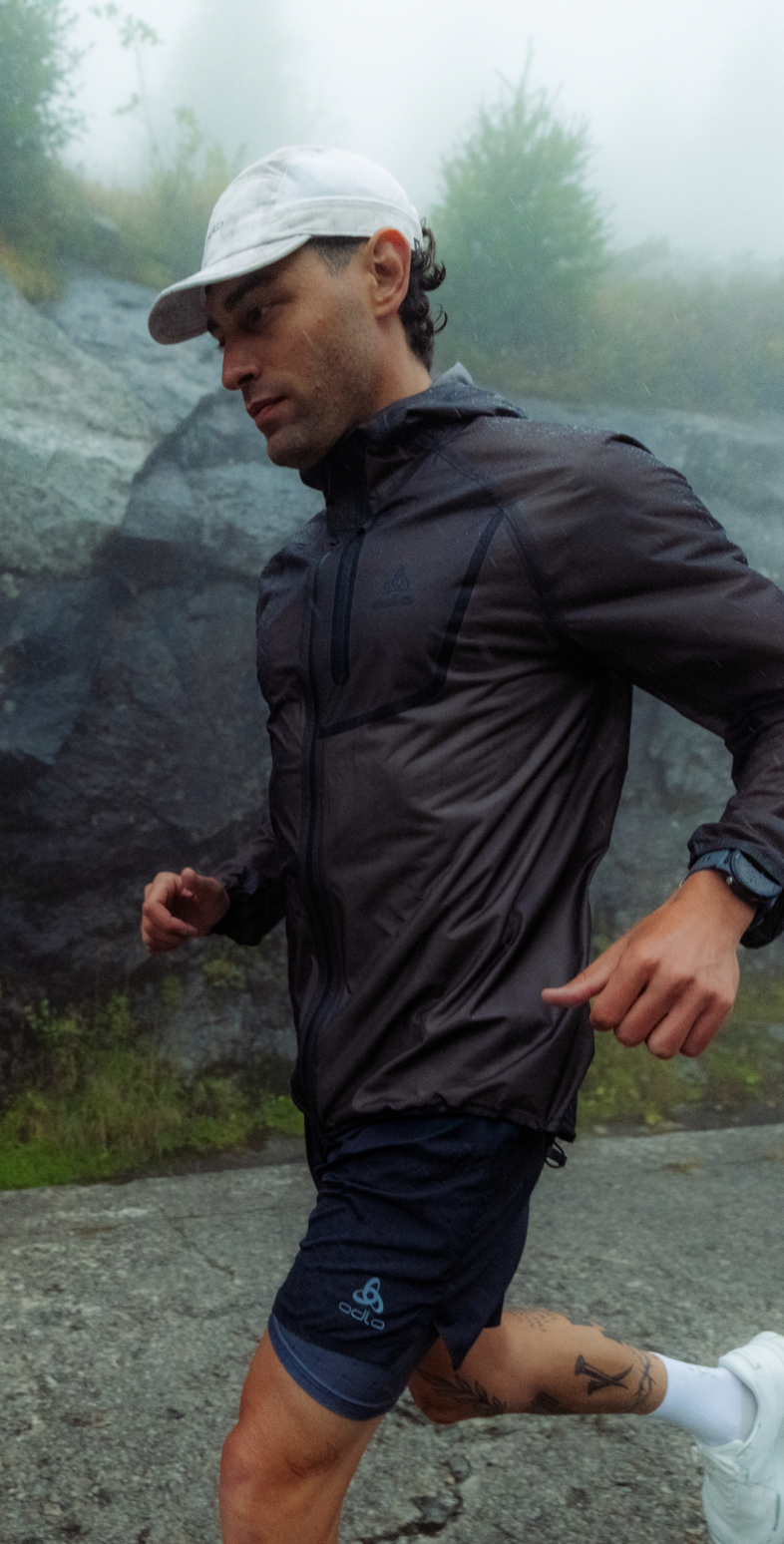 A man running in foggy rain, wearing a white cap, dark rain waterproof jacket, dark shorts, and white trainers on a paved path.