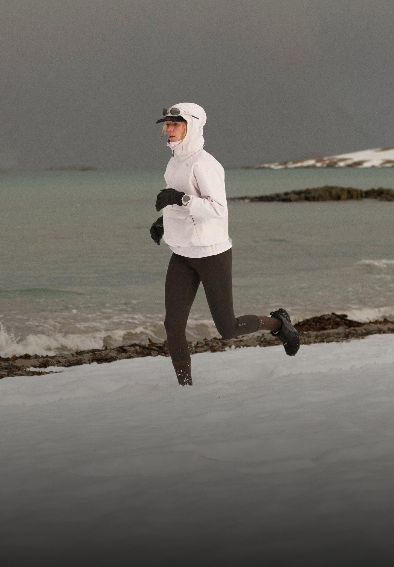 Woman running on a snowy beach wearing a white hooded jacket and black leggings, set against a dark sea and overcast sky.