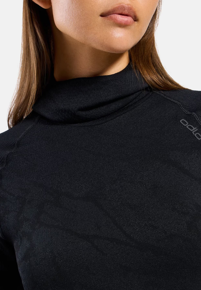 Nahaufnahme einer Frau in einem schwarzen langärmligen Base Layer Oberteil mit Stehkragen und dezentem Logo auf der Schulter vor hellem Hintergrund.