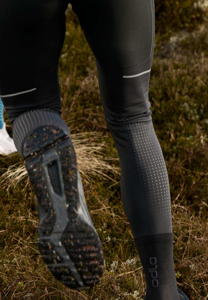 Nahaufnahme der Beine eines Läufers in schwarzen Odlo-Tights mit Reflektionsstreifen, mit einer schmutzigen Schuhsohle auf einem grasbewachsenen Trail.