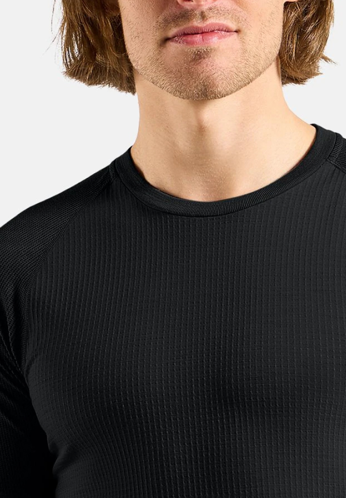 Close-up van het bovenlichaam van een persoon in een aansluitend zwart shirt met textuur, lange mouwen en ronde hals, tegen een lichte achtergrond.