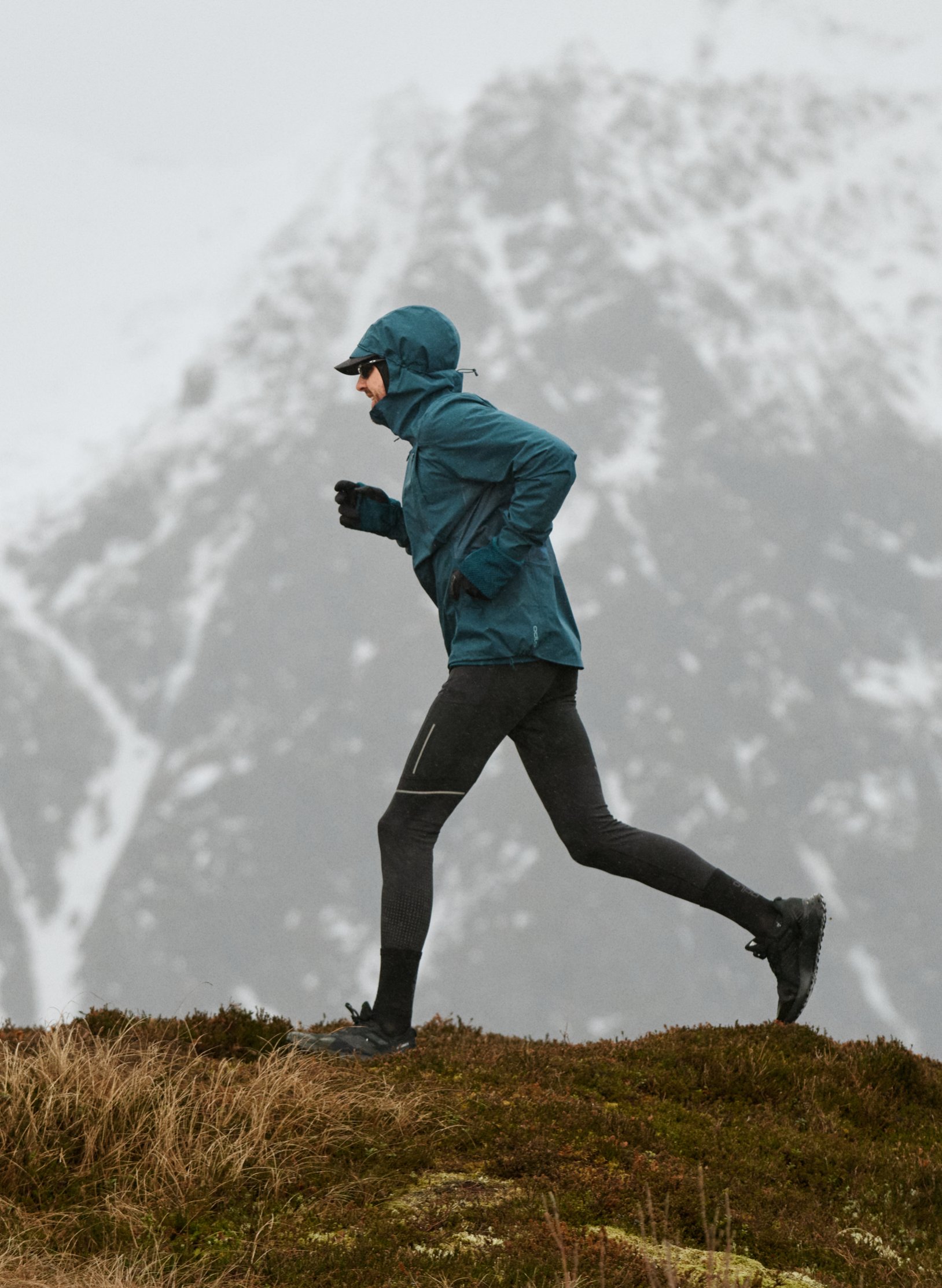 Persoon die trailrunt in een groenblauw jack en zwarte tight op een grasrichel, met op de achtergrond een mistige, besneeuwde berg.