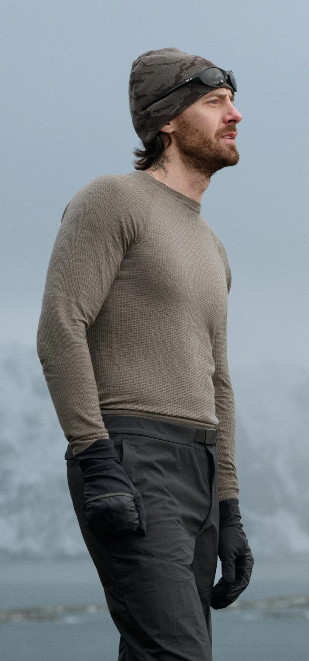 Man and woman in base layers and beanies with sunglasses look into the distance against a foggy mountain backdrop.