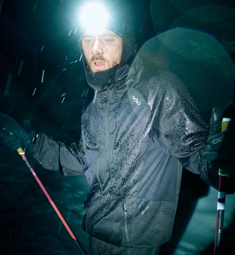 Een man in een natte zwarte jas en met een felle hoofdlamp houdt skistokken vast tijdens een sneeuwstorm in de nacht.