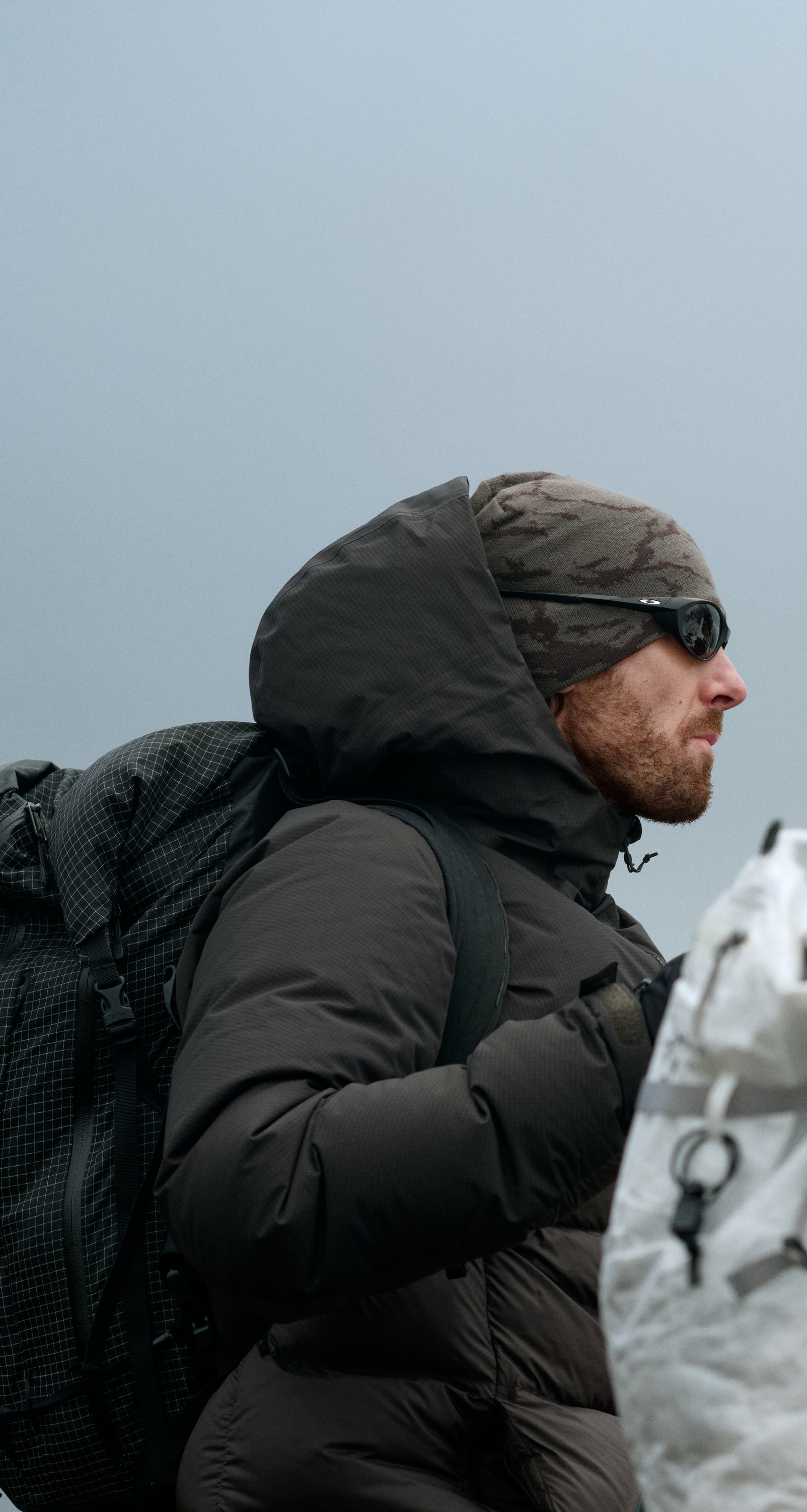 A man in dark winter gear and a woman in a teal jacket, both wearing beanies and carrying backpacks under a grey sky.