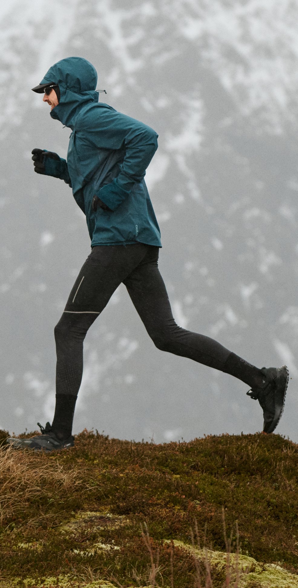 Een persoon in een blauw jack en zwarte legging rent over een grazige bergrug, met besneeuwde bergen op de achtergrond.