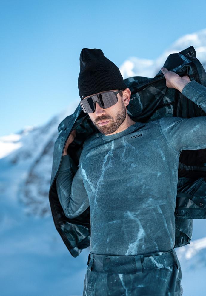 Man in snowy mountains wearing a black beanie, sunglasses, and blue marble-patterned base layer, holding open a jacket.