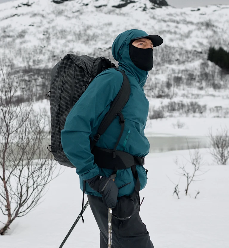 Escursionista con giacca color verde acqua, mascherina nera e zaino, con bastoncini da trekking in mano in un paesaggio invernale innevato e montuoso.