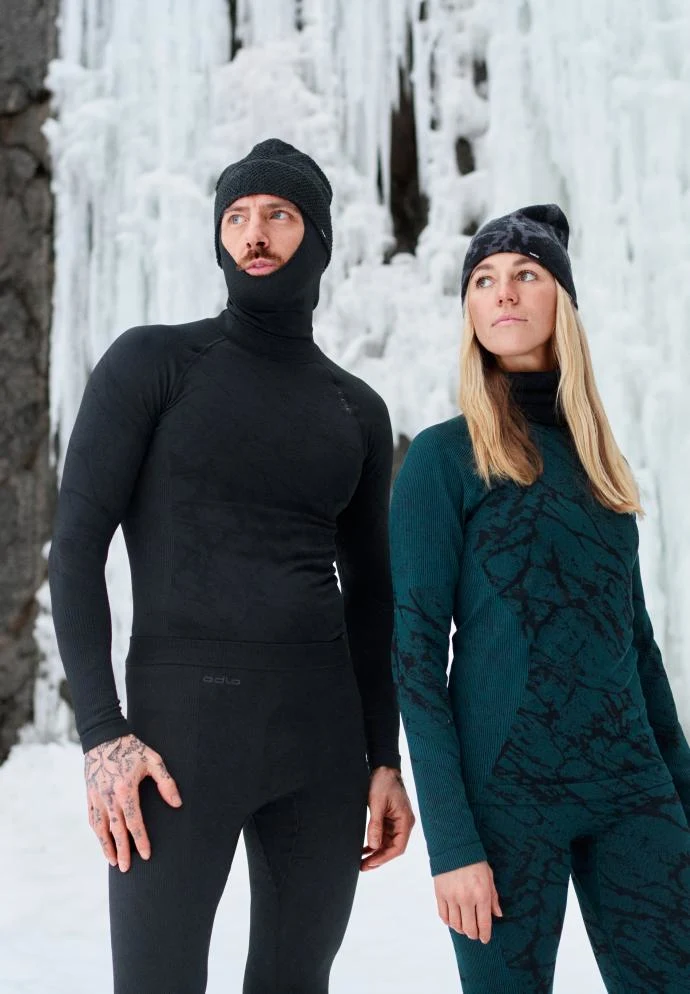 Mann in schwarzer Thermobekleidung mit Kapuze und Frau in türkisfarbener, gemusterter Thermobekleidung stehen im Schnee an einem gefrorenen Wasserfall.