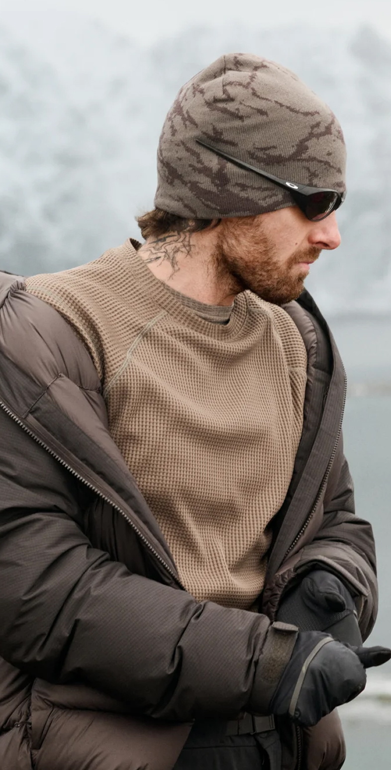 Bearded man with a neck tattoo in a camo beanie and sunglasses looks right, with snowy mountains and water behind him.