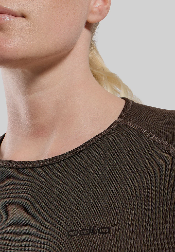 Nahaufnahme eine blonde Frau in einem dunkelbraunen, strukturierten Active Warm Base Layer Oberteil mit Rundhalsausschnitt, dezenten Nähten und dem „odlo“-Logo vor hellgrauem Hintergrund.