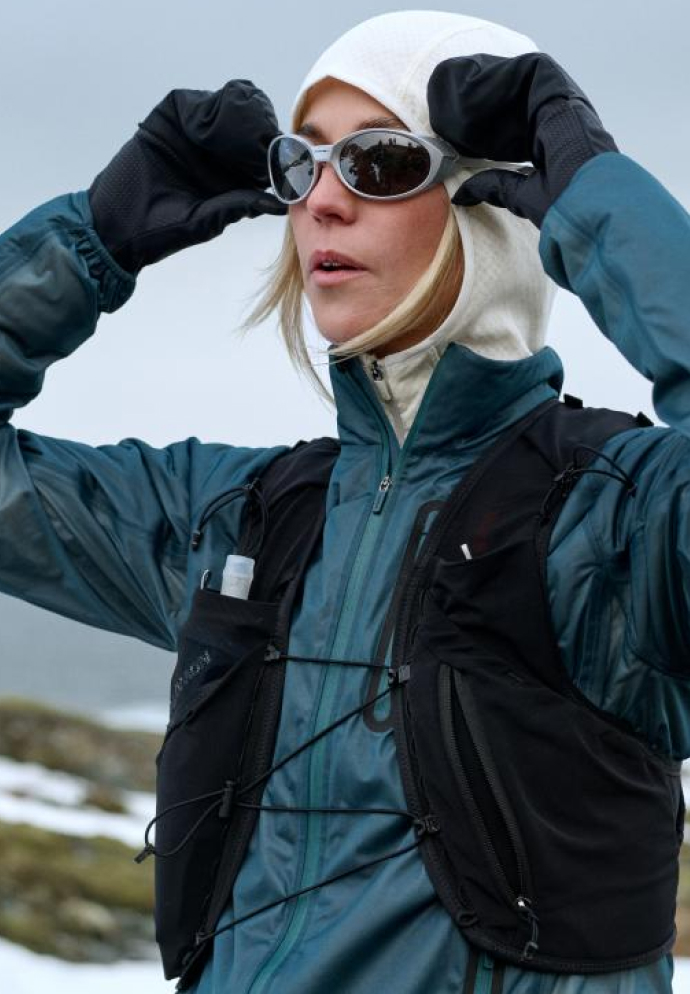 Vrouw in een groenblauw jack, witte capuchon en zwart hardloopdekje past haar zonnebril aan met zwarte handschoenen in een besneeuwd landschap.