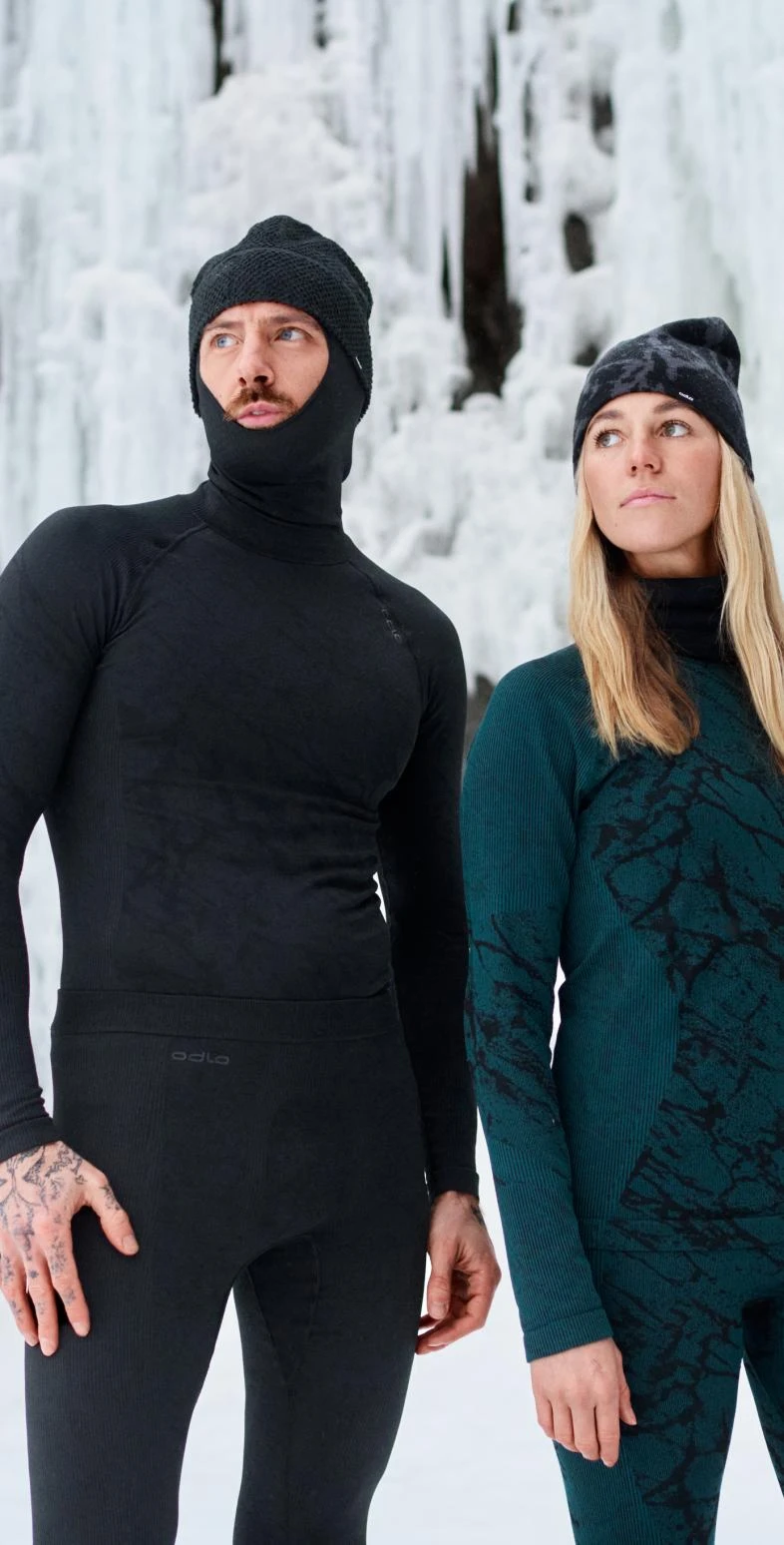 Man in black base layer set and balaclava, woman in green patterned base layer set and beanie, standing against an icy background.