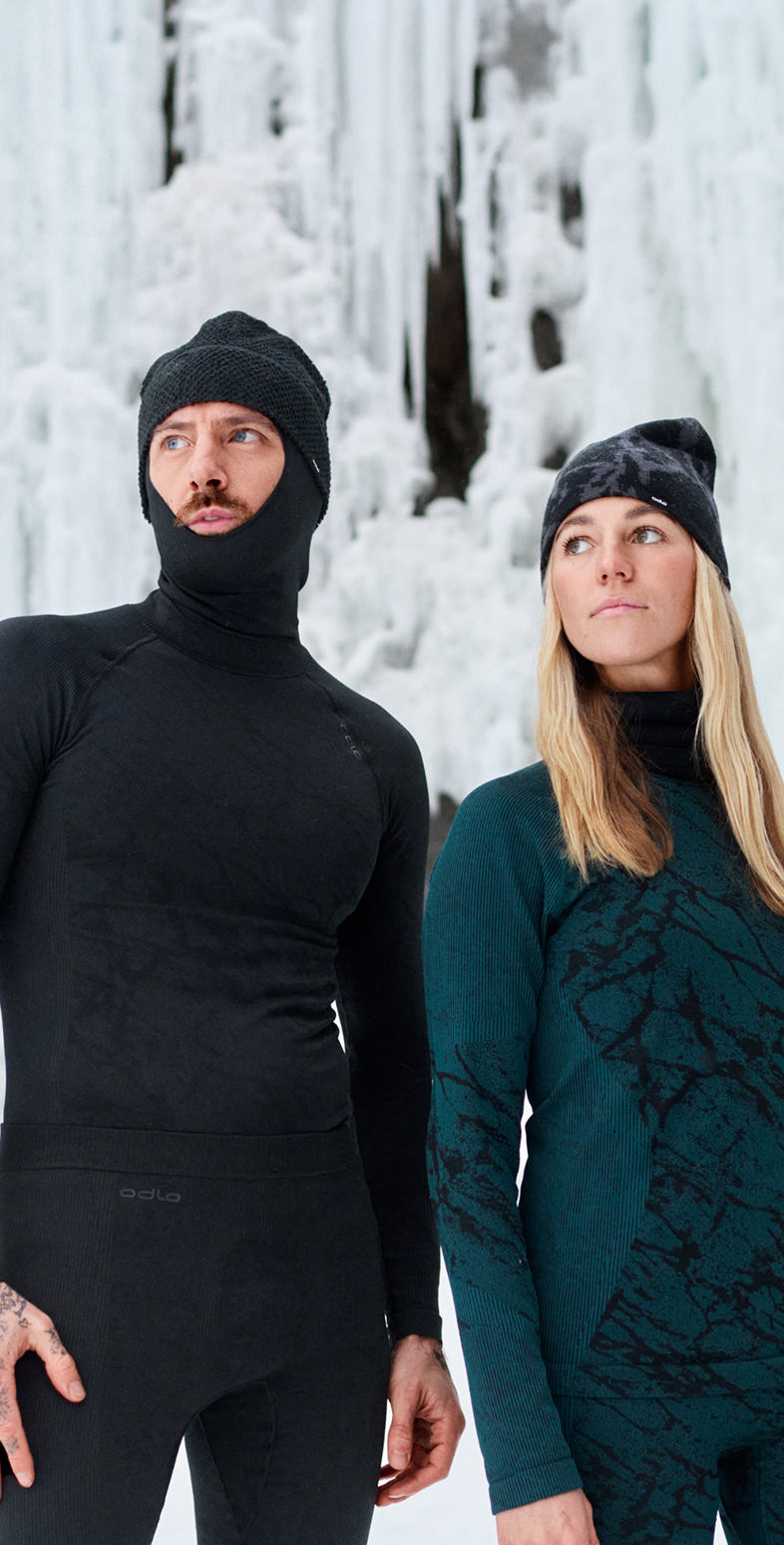 Man in black base layer set and balaclava, woman in green patterned base layer set and beanie, standing against an icy background.