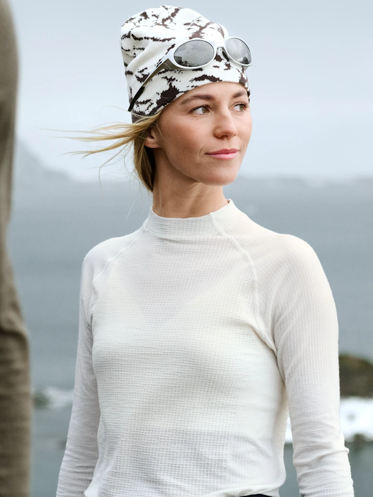 Blonde vrouw in de sneeuw met een donkergroene base layer met zwart marmerpatroon, zwarte beanie en zonnebril.