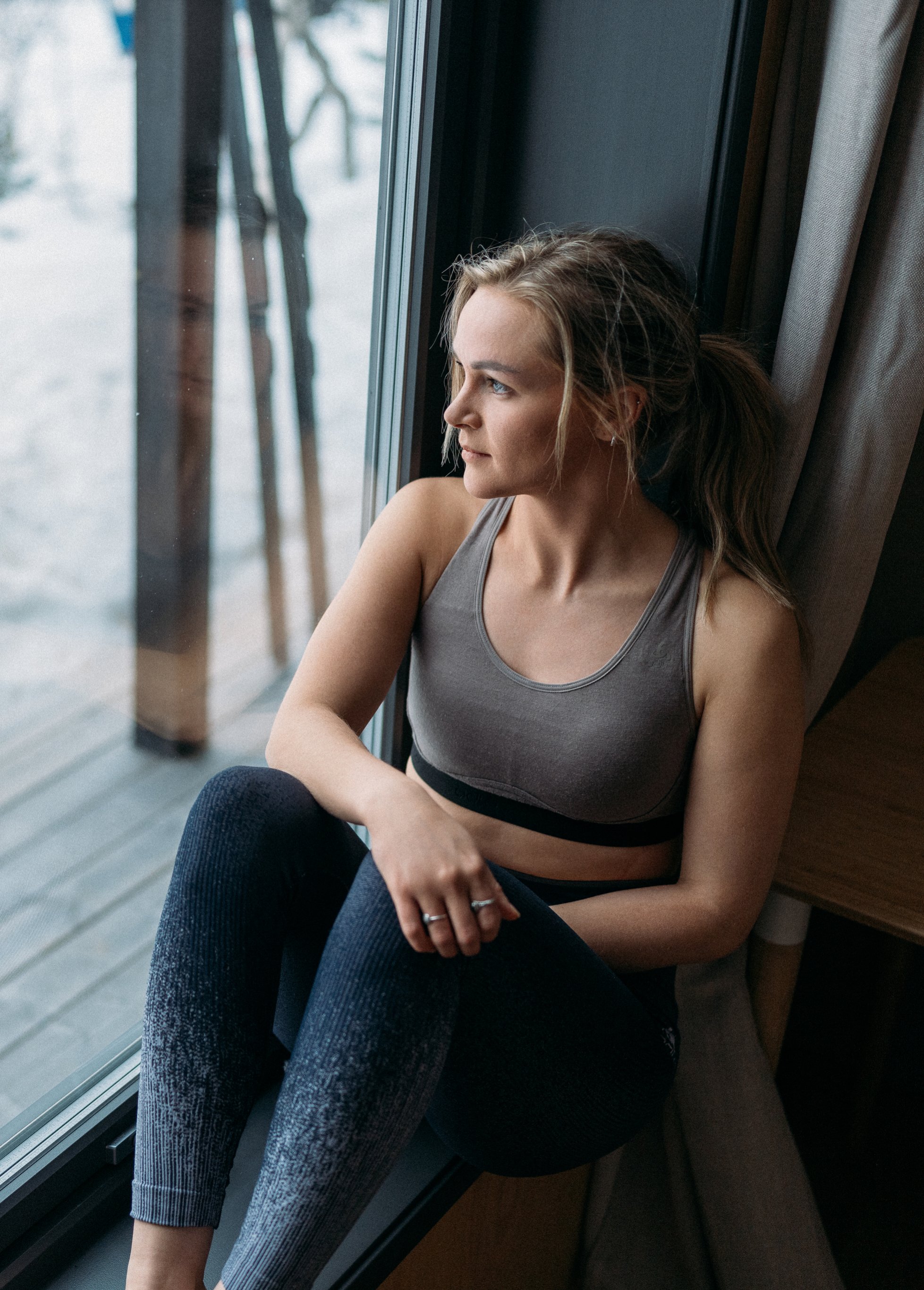 Frau in einem grauen Sport-BH und einer dunklen Leggings sitzt auf einer Fensterbank und blickt nachdenklich auf eine verschneite Landschaft.
