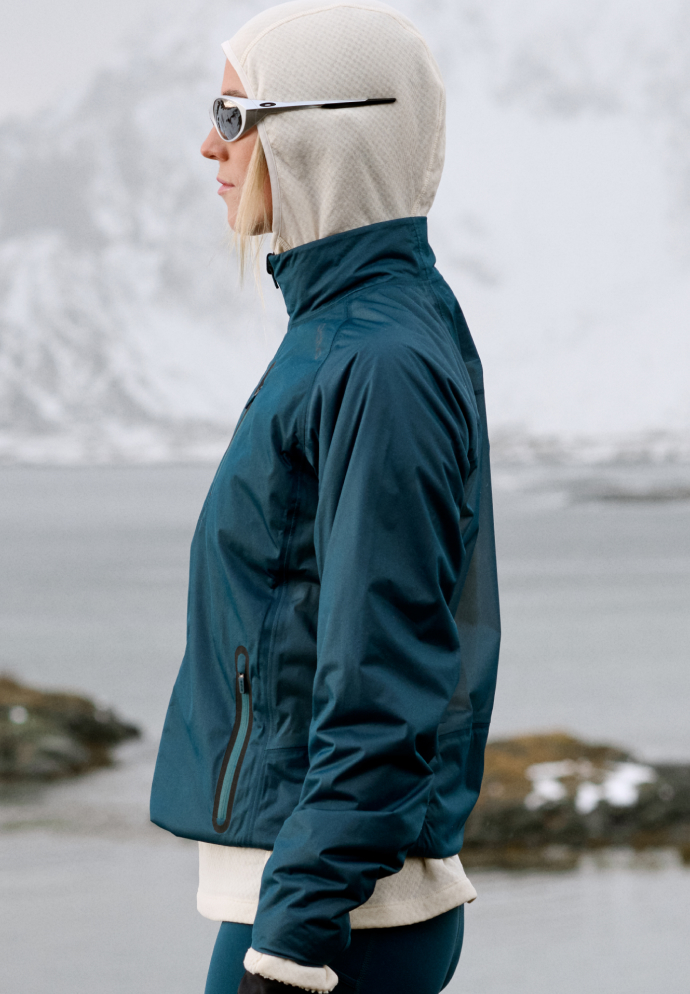 Woman in profile wearing a teal jacket, cream hood and sunglasses, standing by water with snowy mountains behind her.