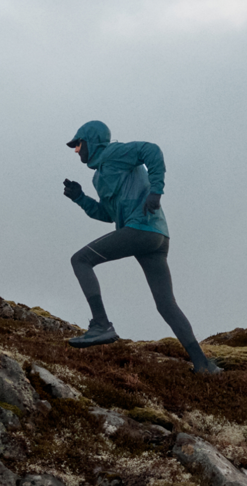 Un runner con una giacca antivento con cappuccio color verde acqua e leggings scuri corre su una collina rocciosa e ricoperta di muschio, sotto un cielo grigio e nuvoloso.